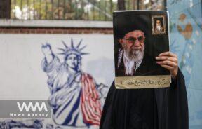 An Iranian woman holds a picture of Iran's Supreme Leader Ayatollah Ali Khamenei next to an anti-U.S. mural on the wall of former U.S. Embassy during the 44th anniversary of the U.S. expulsion from Iran/WANA (West Asia News Agency)