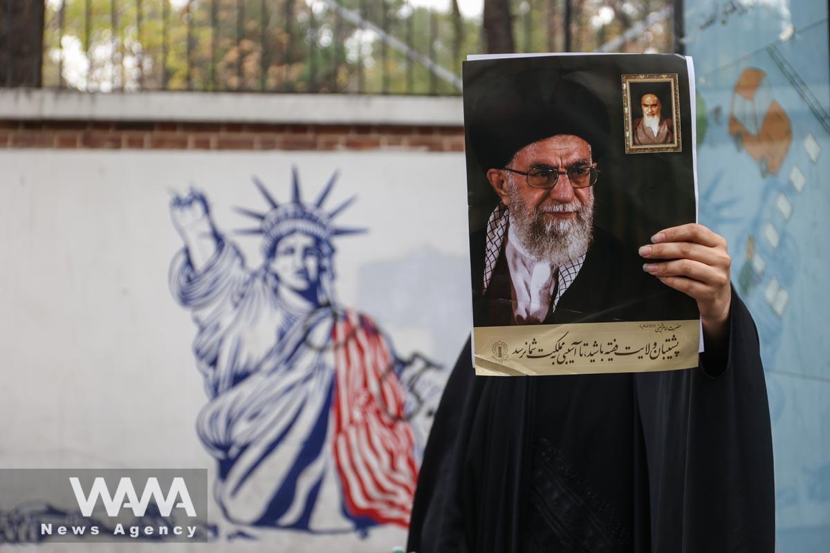 An Iranian woman holds a picture of Iran's Supreme Leader Ayatollah Ali Khamenei next to an anti-U.S. mural on the wall of former U.S. Embassy during the 44th anniversary of the U.S. expulsion from Iran/WANA (West Asia News Agency)