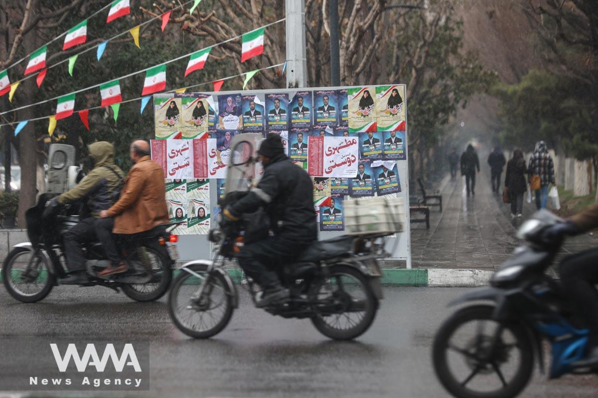 Iranians ride on a motorcycle past campaign posters for the parliamentary election during the last day of election campaigning